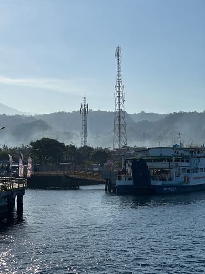 The image shows a ferry docked at a port with two cell towers in the background. The ferry is white and blue, with "We Bridge the Nation" written on its side. A yellow ramp connects the ferry to the dock. In the distance, misty mountains rise behind trees and buildings. The water in the foreground is dark blue and choppy, reflecting the sunlight. The sky is a pale blue with some wispy clouds. The location is likely the port of Ketapang, Indonesia, given the presence of a ferry and the mountainous landscape.