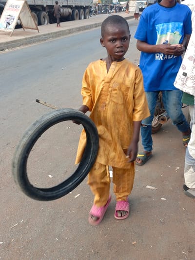 A young boy in a golden-yellow outfit and pink sandals stands on a street holding a tire and a stick. He's near a roadside with other people, including one wearing a blue shirt with "RRADA" on it and another wearing a patterned shirt. A truck and a large sign are visible in the background. The street's surface appears worn and has scattered debris.