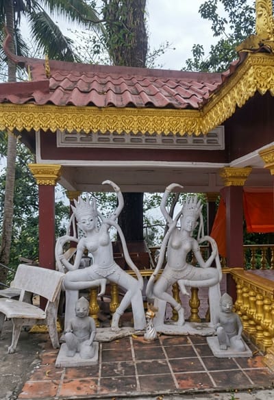 The image depicts an outdoor scene at what appears to be a temple or shrine in Sihanoukville, Cambodia. In the foreground, there are several white stone statues. Two prominent statues are of female figures in dancing poses, adorned with crowns and elaborate headdresses. Between them and slightly to the left is a smaller statue of a seated child or young person. To the right of the dancing figures, another smaller statue of a seated person is visible. A weathered white concrete bench is positioned to the far left. The statues are placed on a tiled floor.
Behind the statues, a structure with a red tiled roof and ornate gold trim is visible, suggesting a religious or ceremonial building. The building features dark red pillars and decorative gold detailing along the eaves and columns. Lush green foliage, including palm fronds and tree branches, surrounds the structure, indicating a tropical or subtropical environment. The sky is overcast. The overall scene is one of cultural and religious artistry set within a natural, outdoor environment.
