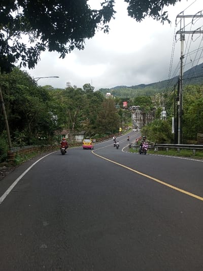 The image displays a curved asphalt road within a hilly, forested environment in Saing Ta Mawt Lay, Myanmar, under an overcast sky. The road features a solid white line on its left edge and a solid yellow centerline. A lone individual on a dark red motorcycle, wearing a red shirt and helmet, travels away from the viewer on the left side of the road, followed by a yellow car with a pink lower rear bumper. Further ahead, a pink scooter with a rider is situated near a metal guardrail on the right shoulder, while several other motorcycles with riders are visible ascending the road. The left roadside is defined by a light brown guardrail and dense green foliage, featuring a streetlight pole. The right roadside includes a metal guardrail, additional green vegetation, and a tall utility pole with numerous lines. The background reveals lush green hills dotted with distant buildings and structures, including what appears to be a bridge or overpass section. The sky is uniformly gray.