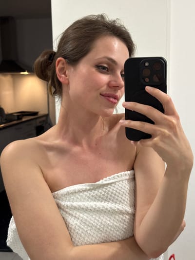 A woman is taking a selfie. She has brown hair pulled back into a bun and is holding a black smartphone with multiple camera lenses up in her right hand. The woman's face is visible, and she is partially wrapped in a white, terrycloth towel. The background shows a white wall and a dark, reflective surface, possibly a mirror, with hints of interior details. The photo appears to have been taken indoors with artificial lighting. There is no information to indicate this image was taken in Kranj, Slovenia.