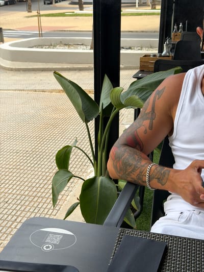 This media file shows a close-up of a person's arm, tattooed with an anchor and other designs, resting on a table next to a plant with large green leaves. The person is wearing a white tank top and a silver bracelet. The background features a textured surface, possibly a table or floor, and a glimpse of an outdoor setting with a pale yellow wall and white planters. The image appears to be taken during the daytime, with natural light casting shadows. There is a QR code visible on a dark gray object in the foreground with the text "NUESTRAS CERVEZAS" and a star icon. The setting suggests a casual outdoor dining or seating area.