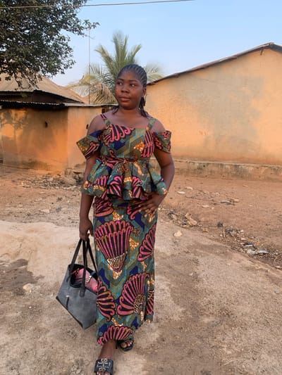 A woman stands outdoors, wearing a colorful, patterned dress with off-the-shoulder details. She holds a large, dark-colored tote bag. The background features a dirt ground and buildings with varied roofing materials, under a bright, clear sky. A palm tree is partially visible to the left of the woman, and other trees are present.