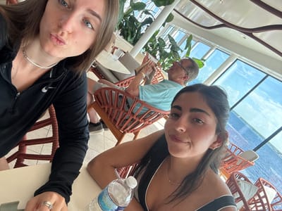 The image captures three individuals within a modern, sunlit establishment featuring expansive windows overlooking a large body of water, consistent with a coastal location in San Miguel de Cozumel, Mexico. In the immediate foreground, a young female with long brown hair and blue eyes wears a black Nike athletic top with a white beaded necklace and a silver ring; she looks directly at the camera with pursed lips. To her right, another young female with long dark hair and dark eyes wears a dark top with white trim and a delicate gold-colored necklace; she smiles subtly at the camera. A clear plastic water bottle rests on the light-colored table in front of her. In the background, an older male with short grey hair and glasses, clad in a light blue collared shirt, occupies a woven chair. He appears focused on an object held in his hands. The environment includes numerous brown woven chairs with light cushions, large green tropical plants in planters, and a contemporary ceiling. Beyond the windows, a vivid blue sky with scattered clouds meets the open blue water.