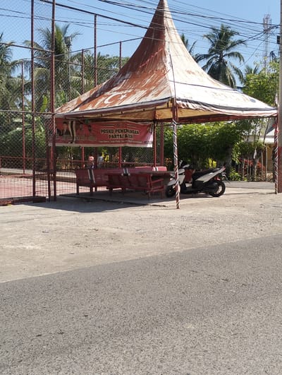 An outdoor scene in Meunasahdayah, Indonesia features a weathered, orange-brown tent with a conical roof over a seating area. Underneath the tent are several red wooden benches and a red wooden table. Behind the tent, a red chain-link fence encloses what appears to be a sports court. A white and black motorcycle is parked to the right of the tent. In the background, lush green trees and palm trees are visible against a bright blue sky. A banner displaying images of men and text in Indonesian hangs on the front of the tent. The ground is a mix of concrete and asphalt.