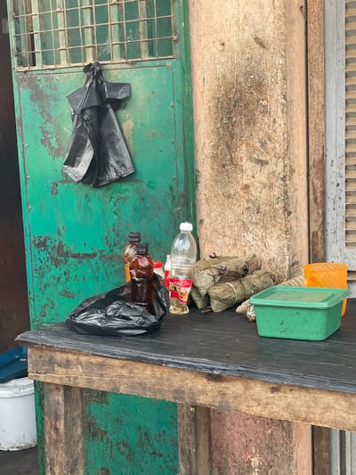 The image displays an assortment of items arranged on and around a weathered wooden surface, suggesting an outdoor or informal setting in Luanda, Angola.
The primary collection of objects rests on a rough, dark wooden plank. On one end, there are two plastic containers: one orange (partially visible) and one green, both rectangular with lids. Beside these are several bundles wrapped in what appear to be plant leaves, tied with string, resembling traditional food preparations like tamales. Further along the plank, a clear plastic bottle with a red and yellow label is visible, next to a brown plastic bottle that is partially covered by a black plastic bag. Another crumpled black plastic bag, containing more brown bottles, lies nearby.
The wooden plank is positioned over a green, metallic surface that shows significant signs of rust and wear, possibly a large generator, utility box, or a sturdy metal table. This green surface also features a rectangular metal grate on one side. A crumpled black plastic bag is also present on this green surface. The background of the scene is a rough, light brown or grey concrete wall. Below the wooden plank, part of a white bucket with a blue handle can be seen.
The arrangement of objects suggests they could be goods laid out for sale at an informal market stall or simply items in temporary storage. No people are visible, and no specific activity is actively taking place. The overall impression is one of a functional, somewhat rustic outdoor space. The items and surfaces are worn, reflecting exposure to the elements. No clear, legible text is visible on any of the items or surfaces. The lighting indicates it is daytime.