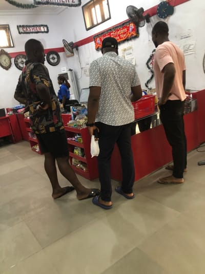 Three men lean against a counter in what appears to be a store. The first man, wearing a pink shirt and black pants, leans on the counter. The second man, in a patterned shirt and black pants, rests similarly. The third man, wearing a patterned shirt and dark shorts, is barefooted. Red shelves are behind them filled with various items. The store also features Christmas decorations, signs labeled "Cashier", and decorative clocks on the walls.