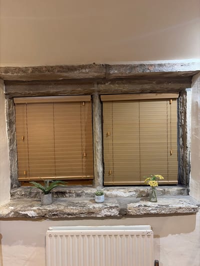 The image shows a close-up interior view of a window recess with two wooden blinds. The window is framed by rough-hewn stone. On the stone windowsill below the blinds, there are two small potted plants and a small glass vase with yellow flowers. Below the windowsill, a white radiator is visible. The overall impression is of a rustic interior. The image location is not specified in the provided context.