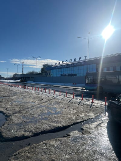 This image depicts the exterior of Murmansk Airport in Murmashi, Russia, under a clear blue sky with bright sunlight, causing a lens flare in the upper right quadrant. The primary subject is the airport terminal building, which features a modern design with large, blue-tinted reflective glass windows across its facade. Above these windows, large white letters spell "АЗРОПОРТ МУРМАНСК - СТОЛИЦА АРКТИКИ" (AIRPORT MURMANSK - CAPITAL OF THE ARCTIC). To the left of the main terminal, a multi-level structure resembling a control tower is visible, partially obscured by scaffolding, indicating ongoing construction or maintenance.
The foreground is characterized by a patchy, uneven terrain of melting snow and ice, mixed with dirt and numerous puddles of water, suggesting a thaw. A distinct crack or channel, filled with water, runs across the immediate foreground. A line of approximately 15-20 flexible white bollards with orange reflective stripes separates this slushy area from a paved road surface. On the road in front of the terminal, a dark-colored passenger bus with a light blue and purple gradient design on its side is visible. The front right fender and wheel of a dark-colored car are partially visible in the extreme lower right corner of the frame. Multiple streetlights with dual luminaires are positioned along the road leading towards the terminal. No individuals are discernible within the scene.