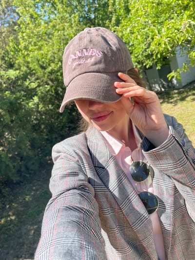 A person wearing a plaid jacket and a baseball cap is standing outdoors, likely in a sunny area, as indicated by the hand shielding the eyes. Sunglasses are visible on the jacket. The background appears to be a green, leafy area. The person is wearing a pink top under the jacket. There is no evidence to place the location of the image in Indjija Municipality, Serbia.