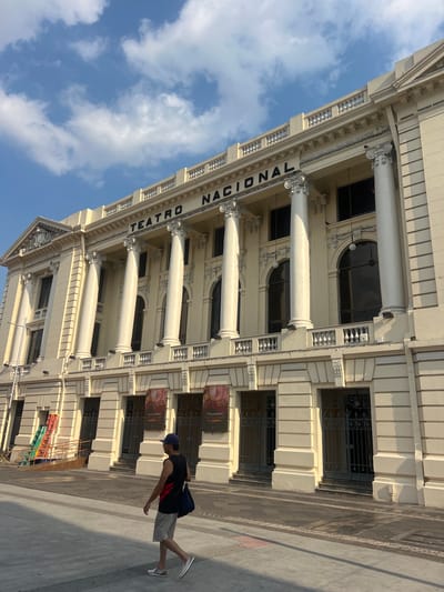 The image shows a slightly tilted view of the Teatro Nacional, located in San Salvador, El Salvador. The neoclassical building features cream-colored stone, columns, arched windows, and decorative molding. The words "TEATRO NACIONAL" are vertically inscribed in capital letters on the facade. In the foreground, a man wearing a black tank top, grey shorts, a blue cap, and white sneakers is walking past the building. A blue bag hangs from his shoulder. The sky above the building is blue with scattered white clouds. Some construction equipment is visible at the bottom of the frame, along with a poster on the facade.