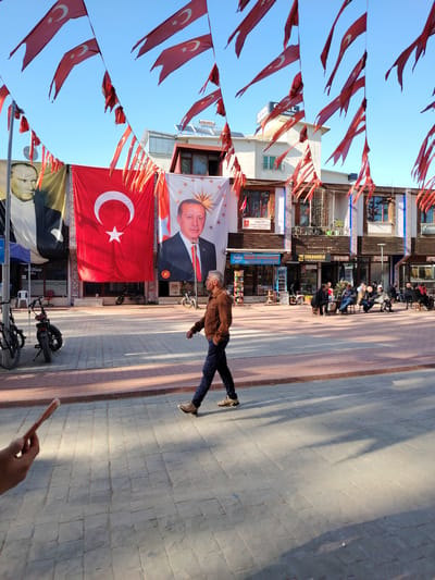 This street-level image captures a bustling scene in Iznik, Turkey, filled with nationalistic displays under a clear blue sky. Numerous Turkish flags, featuring the white star and crescent on a red background, are strung across the street, many appearing slightly worn.

Three prominent banners adorn the building facades in the background: a portrait of Mustafa Kemal Atatürk on the left, a large vertical Turkish flag in the center, and a portrait of President Recep Tayyip Erdoğan on the right.

In the foreground, a man in a brown jacket and dark trousers walks across a light-colored paved square, his shadow stretching long on the ground, indicating either mid-morning or late afternoon sun. A hand holding a mobile phone is visible in the bottom-left corner. On the right side of the square, several men are gathered at small tables outdoors, suggesting a cafe or social gathering spot. Parked motorbikes are visible on the left.

The buildings house various businesses, with legible signs including "ERKANOĞLU GAYRİMENKUL" (real estate), "Gay Ocağı" (tea house/cafe), "DERİZ KURUYEM" (dried nuts/fruits), "OTEL RİMEDY" (hotel), and "ATSAN." Other visible text includes "TÜRKİYE" and "ZAB MİNÖKTASI" (likely "Zabıta Noktası" - municipal police point). The scene conveys a sense of everyday life amidst a strong display of national pride.