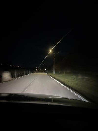 The image depicts a night scene from the perspective of a vehicle's interior, looking forward through the windshield onto a road in Carrazeda de Ansiães, Portugal. The sky is dark, indicating nighttime. The vehicle's headlights illuminate the asphalt road ahead, which features visible white lane markings and a central dashed line. The lower portion of the image shows the dark top edge of the vehicle's dashboard or windshield frame.
On the right side of the road, a bright street light mounted on a utility pole is prominent, casting a distinct starburst effect. Further along the right side, another utility pole is visible, along with green vegetation that appears blurred due to motion, suggesting the vehicle is moving. A round white sign is also visible on the right, further down the road.
On the left side of the road, a light-colored retaining wall or barrier runs parallel to the road, also showing slight motion blur. Beyond this, a darker area of vegetation or landscape is present. Distant lights are faintly visible on the horizon, suggesting structures or lights further away. No individuals are visible in the scene. The overall impression is a vehicle traversing a rural or semi-urban road at night.