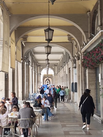 A long colonnade, likely in Cagliari, Italy, features a succession of arches. Hanging lamps descend from the arched ceilings. A mix of seated customers at outdoor cafes and pedestrians occupies the shaded walkway. Some people are sitting and eating while others are walking through the corridor. A woman in black clothing walks away from the viewer. Flowers hang above the entrance on the right side of the image.