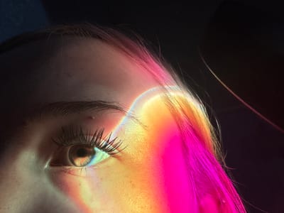 This media file captures a close-up, artistic shot of a person's eye and hair, illuminated by colorful light reflections. The primary subject is the eye, with its eyelashes and iris clearly visible. The light creates a rainbow effect across the eye and part of the person's face, casting hues of pink, yellow, and orange. The person's hair, colored in vibrant pink and blonde tones, is also prominently featured, catching the light and blending into the colorful reflections. The background is dark and abstract, with some hints of texture suggesting a fabric or a soft surface.
The activity is a still image, likely a portrait or conceptual photography. There is no discernible event or action taking place, but the composition suggests a deliberate artistic arrangement. The notable details include the striking color play from the light, the sharp focus on the eye and eyelashes, and the soft diffusion of light on the skin and hair. The lighting implies it might be artificial, as it creates distinct, sharp color bands, but the overall mood is somewhat ethereal and dreamlike. There is no visible text in the image. The location context of Velykyi Sukhodil, Ukraine, does not provide any direct visual cues within the image itself.