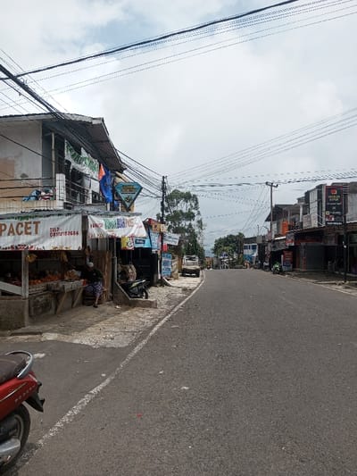A daytime scene depicts a street lined with buildings, shops, and power lines. A fruit stand is visible on the left with a person seated. A red scooter is parked on the left side of the road. A silver truck is parked further up the road, with scattered motorcycles and cars visible along the street. The sky is overcast. The road pavement is visible and extends to the distance.