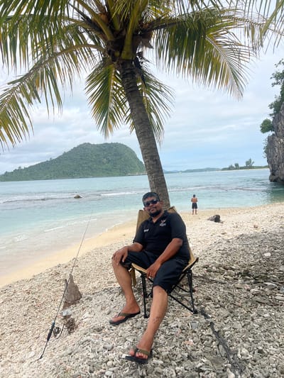 The image depicts a tropical beach scene. The primary subject is a man sitting on a folding chair on a rocky shore. He is wearing a black t-shirt and black shorts, sunglasses, and flip-flops. To his left, a palm tree with large fronds dominates the foreground. In the background, the calm turquoise ocean meets a sandy beach, backed by a lush, green, mountainous island. Another person is visible in the shallow water, standing near the shore. A fishing rod is set up on the beach, suggesting fishing is taking place. The sky is overcast, indicating cloudy weather. The overall mood is relaxed and vacation-like, characteristic of a beach setting. The time of day appears to be daytime, given the lighting. No visible text is present. The location context provided is Kruengtunong, Indonesia, which aligns with the tropical scenery.