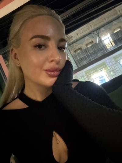 A woman with blonde hair and fair skin is angled in a selfie. She wears a black top with a triangular cutout revealing a chain necklace. Outside the window behind her are ornate, tall buildings. The background is softly illuminated, while the woman's face is in focus, highlighting her makeup and facial features. The shot is taken from a low angle, emphasizing her upward gaze.