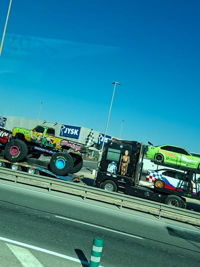 A flatbed truck carries several vehicles on a road in San Fulgencio, Spain. On the truck's trailer, a lime green monster truck with elaborate monkey-themed artwork and the word "Monkey" written on the side is positioned on the left. In front of the monster truck, the side of a red vehicle is partially visible. To the right of the monster truck, a black truck is carrying two cars on its upper deck. The top car is a lime green sedan with racing decals and the text "Cristian Motor Show" and "Power". Below it, a white, red, and blue checkered car with orange rims is visible. The truck and its cargo are positioned on a multi-lane road with a guardrail. In the background, a long, white building with a blue "JYSK" logo is visible, along with streetlights and power lines. The sky is clear and blue.