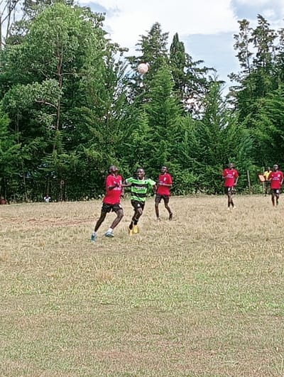 An outdoor sports scene in Lwandeti ward, Kenya, depicts six male individuals on a dry grass field backed by a dense line of tall green trees and a partly cloudy sky. A light-colored ball is airborne in the upper center of the frame. In the foreground, two players are in motion: one on the left wears a red shirt, dark shorts, and blue and white athletic shoes, while another in the center wears a green and black striped shirt, dark shorts, and yellow athletic shoes. Three additional players, all in red shirts and dark shorts, are visible in the mid-ground, moving towards the right side of the frame.