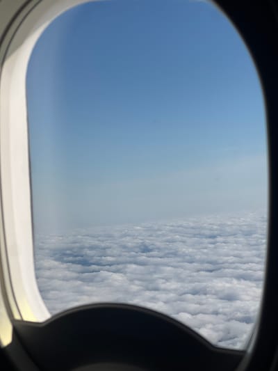 An airplane window frames a view of a vast expanse of white clouds stretching to the horizon under a clear blue sky. The window frame is dark on the right side and white on the left. The clouds appear fluffy and textured, suggesting they are at a significant altitude. No city or specific geographical features are visible, making it impossible to ascertain a location in Kczewo, Poland. The scene depicts a typical view from an airplane in flight.