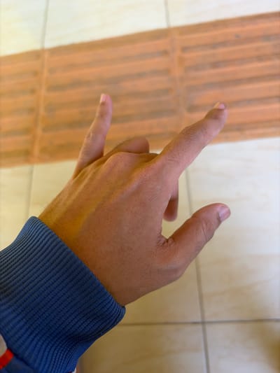 A close-up, overhead shot shows a person's right hand performing the "rock on" or "horns" gesture. The hand is positioned in the center of the frame, with the index finger and pinky finger extended upwards, while the middle and ring fingers are curled downwards and held by the thumb. The hand is wearing a dark blue ribbed cuff, likely from a long-sleeved garment. The background consists of a tiled floor, with a textured, orange section on the left half and a lighter, smoother tiled section on the right. The lighting suggests an indoor environment. This image was captured in Pasekaran, Indonesia.