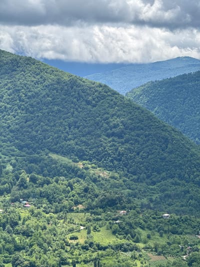The image depicts a mountainous landscape in the city of Gagulechi, Georgia. The foreground is dominated by lush green forests covering the slopes of hills and mountains. Several small houses with red and white roofs are scattered throughout the lower part of the image, nestled among the trees. Winding paths and agricultural terraces are visible on the hillsides. In the background, layers of further mountains recede into the distance, appearing progressively bluer due to atmospheric perspective. The sky is filled with a mix of dark, dramatic clouds and brighter, overcast sections. The overall scene conveys a sense of rural tranquility and natural beauty.