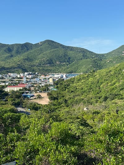 The image depicts a panoramic view of a coastal town nestled between lush green mountains and a clear blue sky. In the foreground, dense foliage covers the lower portion of the frame, with a winding road visible amidst the greenery. Further into the scene, a cluster of buildings with varying architectural styles forms the town. These buildings are mostly white with some featuring red roofs, suggesting a residential or commercial area. Among them, a prominent building with a yellow facade and a red-tiled roof stands out. Beyond the town, a body of water, likely the sea, is visible with several boats anchored, indicating a port or harbor. The background is dominated by rolling green mountains, their slopes covered in thick vegetation. The overall impression is a serene and picturesque landscape, possibly a tourist destination. The location is identified as Vinh Hai, Vietnam, a coastal commune known for its natural beauty and fishing communities. The scene appears to be captured during daylight, with bright sunlight casting shadows and illuminating the landscape.