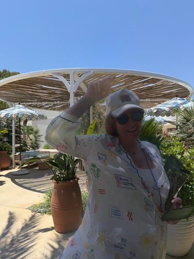 A woman is pictured outdoors, wearing a white cap with a logo, sunglasses, and a white patterned shirt. She is smiling and waving her hand. The woman is seated near a large, circular structure with a thatched roof, possibly an umbrella or part of a canopy, and is surrounded by greenery. A terracotta pot with a plant is visible beside her, and palm tree shadows are cast on the ground. The scene appears to be in a sunny, outdoor location, likely during the day given the bright light and clear blue sky. The pattern on her shirt features small illustrations of items like sardines, waves, and crabs, along with text elements such as "Sardines" and "Lemon Oil." The overall atmosphere suggests a relaxed, possibly vacation setting. The provided location context places this scene in Pyrgos Community, Cyprus.