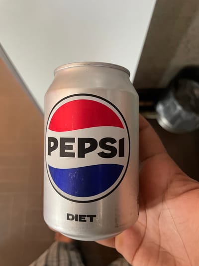 A silver aluminum can of Diet Pepsi is held in a person's right hand. The can features the classic Pepsi logo with red and blue colors within a white circle, and the word "PEPSI" is prominently displayed in black text. Below the logo, the word "DIET" is written in black capital letters. The background is blurred, showing a light-colored surface, possibly a wall or countertop, and a darker area that could be furniture or another object. The lighting suggests an indoor environment. No specific city or geographical location is discernible from the image itself.
