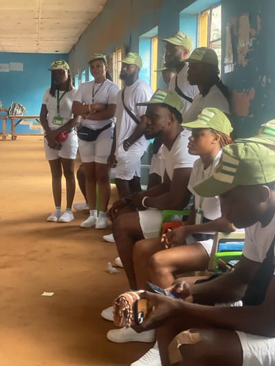 In Ise-Ekiti, Nigeria, a group of young people, likely participants in a youth service program given their matching white t-shirts, white shorts, and green caps with a white emblem, are gathered indoors. The room appears to be a classroom or hall with light blue painted walls and a textured ceiling. Natural light streams in from windows on the right side of the frame, illuminating dust particles in the air. The floor is a reddish-brown, possibly compacted earth or dirt. Several individuals are seated on colorful plastic chairs, while others stand. Some are looking at their mobile phones, suggesting a moment of downtime. One person in the foreground is holding a phone with a decorative phone case. Another individual is seen with a bandage on their knee. The overall atmosphere seems relaxed, with the young people in a communal setting.