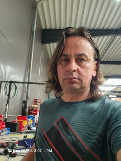 The image is a medium shot, taken from a slightly low angle, of a man with medium-length brown hair and light brown eyes. He is wearing a dark teal t-shirt with a graphic design. The man is positioned in the foreground, looking directly at the camera. The background appears to be an indoor industrial or workshop setting, with visible shelving units filled with red and yellow bins, tools, and equipment. The lighting is bright, suggesting artificial lighting common in such environments. A timestamp in the lower left corner of the image reads "30 [June] 2026 3:07 PM". While the image itself does not provide explicit geographic markers for Papenburg, Germany, the presence of industrial equipment and the overall aesthetic suggest a setting that could be found in a manufacturing or storage facility within an urban or industrial area.