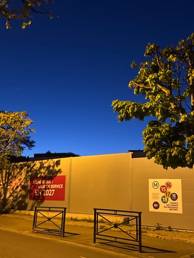 The image captures an urban night scene, predominantly featuring a deep blue sky and elements of a street and building. The perspective looks upwards, showing the dark sky at the top and left, partially obscured by illuminated tree foliage. On the right side, a light-colored building wall stands vertically, with a paved road or sidewalk at its base.
The time of day is clearly night, evidenced by the dark blue sky and the artificial yellow light that illuminates the tree leaves, the building wall, and the ground, casting distinct shadows of trees onto the wall and pavement. Two dark, square metal barriers are visible on the paved surface, positioned in front of the wall.
Two large rectangular posters are affixed to the building wall. The upper poster, predominantly white, features a large "M" logo and several colored circles with the numbers "15", "16", "17", and "18", indicating different metro lines. Adjacent to these, text in French reads "SUIVEZ LES LIGNES DU NOUVEAU MÉTRO!" (Follow the lines of the new metro!). The lower poster is red with white text, stating "LIGNE 15 SUD: MISE EN SERVICE EN 2027" (Line 15 South: put into service in 2027). These posters indicate ongoing or upcoming public transport developments, specifically related to the Grand Paris Express in the Champigny-sur-Marne area. There are no people visible, and no active events are taking place, only static urban infrastructure and promotional material.