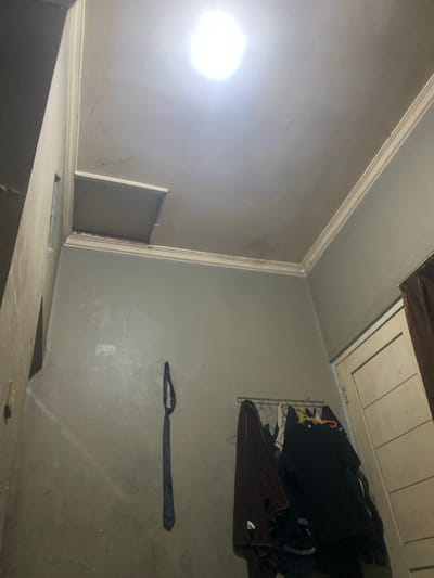 The image shows a corner of a room in Purwokerto, Indonesia, with a white ceiling and gray walls. A light fixture is mounted in the ceiling, illuminating the room. The ceiling is cracked and has water stains. A dark tie hangs on the wall to the left. On the right wall, a metal rod hangs from the wall, and several clothes hang from it. A door is partially visible on the right, with a brown curtain behind it.