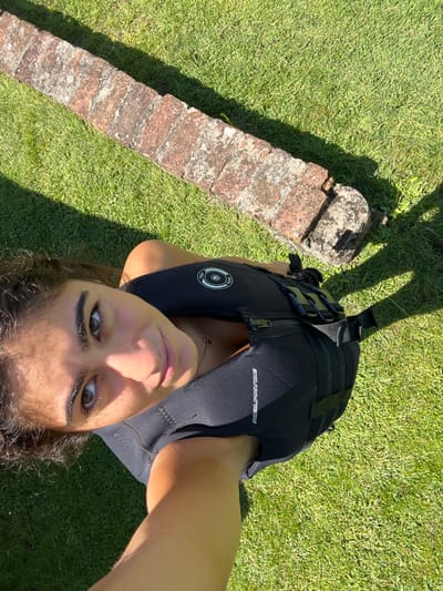 A female subject, appearing to be a young adult, is depicted outdoors in Coruche, Portugal. She is wearing a black NeilPryde brand flotation vest or impact vest, featuring a front zipper, adjustable straps with buckles on the side, and a circular emblem on the upper chest. Her dark, curly hair frames her face, and her brown eyes are directed towards the camera in a self-portrait. The background consists of vibrant green grass and a low, linear structure made of weathered reddish-brown bricks, partially covered in light-colored lichen or moss. Bright sunlight illuminates the scene, casting distinct shadows on the grass from both the subject and the brick structure.