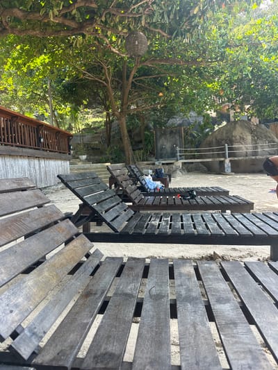 The image captures an outdoor, sunny scene, likely a relaxation or dining area, set amidst natural surroundings. The main subjects are numerous dark wooden benches and tables, arranged on a light-colored ground surface, possibly sand or concrete. Some of these wooden structures appear to be built against a low wall or raised platform.
To the left, a dense canopy of lush green trees dominates the midground, with bright sunlight filtering through their leaves, creating dappled shadows. A decorative, spherical, woven lamp or lantern hangs from a branch of one of these trees. Further into the background, a substantial natural rock formation or boulder is visible, partially obscured by foliage.
On the right side of the image, the lower body and legs of a person are partially visible. Near the center, a simple clothesline is strung between posts, with what appear to be clothes or towels hanging to dry. On one of the tables or benches beneath the clothesline, a few items, possibly clothing or bags, are piled. A wooden deck or walkway with railings is visible in the lower-left background, winding through the greenery.
The scene suggests a relaxed atmosphere in a natural, possibly tropical, setting, consistent with the provided location context of Koh Tao, Thailand. The bright sunlight indicates it is daytime, and the weather appears clear and pleasant. There is no specific activity overtly taking place, but the setup implies a space for leisure or casual dining. No visible text is discernible in the image.