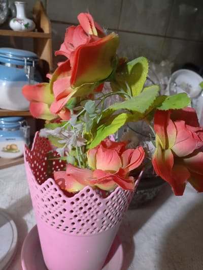 The image depicts a static interior scene in Nasiriyah, Iraq, focusing on a floral arrangement in a domestic setting.
In the foreground, a pink cylindrical pot with a decorative perforated rim, possibly made of plastic or ceramic, holds an arrangement of artificial flowers. The flowers are primarily fabric, featuring coral-pink petals with yellow-green bases, resembling roses, interspersed with smaller sprigs of lighter green and white artificial foliage. The pot rests on a matching pink saucer, both positioned on a white, textured surface, likely a tablecloth or countertop.
The background on the left reveals a wooden shelf. On this shelf, a small white ceramic vase with painted floral details is visible. Below the shelf, a blue and white ceramic or glass canister with a lid is present. This canister bears a label that reads "Spices" and shows an illustration of two small, light brown, oval-shaped objects. A second, similar blue and white canister is partially visible beneath the first.
The background on the right side of the image features a dark-colored wall, possibly tiled. Several out-of-focus objects, appearing to be white plates and clear glassware, are discernible on a surface, suggesting a kitchen or dining area. The lighting appears to be artificial. No individuals are present in the scene, and no discernible actions or interactions are occurring.