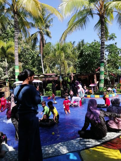 The image depicts a lively outdoor swimming pool scene in what appears to be a water park. The pool has bright blue water and is populated with numerous individuals, predominantly children, engaged in various water activities. Several children are splashing, playing, and swimming, with some using inflatable rings. Adults are also present, supervising and participating.
The environment is tropical, characterized by lush green vegetation and tall palm trees that cast shadows across the scene. In the background, there are structures that suggest amenities like a covered seating area or changing rooms, decorated with colorful, whimsical mushroom-shaped elements. The ground surrounding the pool is a mosaic of colorful tiles in shades of blue, yellow, and red. The sky is clear and blue, indicating a sunny day.
While the image doesn't explicitly state the location as Tegalwangi, Indonesia, the presence of tropical flora, local attire on some individuals (such as headscarves), and the general aesthetic are consistent with such a setting in Indonesia. The overall impression is one of a fun and recreational family outing at a water park.