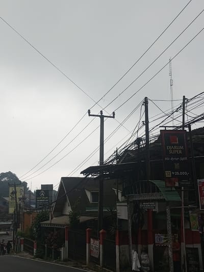 An overcast street scene showcases a dense network of overhead power lines against a grey sky. A tall telecommunications tower pierces the horizon. Buildings are visible, including one with a gabled roof. A "Djarum Super" advertisement hangs prominently, with price information in Indonesian Rupiah. Street signs and political posters add visual clutter. A fence in red and white stripes runs along the roadside. The image overall appears to be taken in a densely populated area in Indonesia.