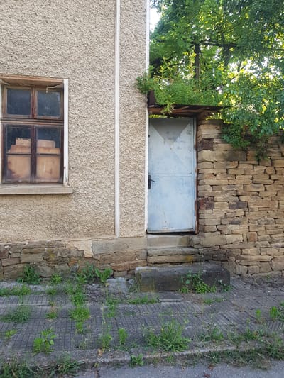 An exterior shot captures a weathered building facade in Lomets, Bulgaria. A single, light-blue metal door is set into the building, flanked by a rough-hewn stone wall on the right and a textured, pale stucco wall on the left. The door has a black handle and appears to be slightly ajar, with a small, weathered metal overhang above it. To the left of the door, a multi-paned wooden window with dirty glass offers a glimpse of objects stacked inside. The building's base is constructed with rough stones, and the ground in front is a gray, grid-patterned paved area interspersed with weeds and grass. A large, dark gray concrete block sits to the right of the doorway. Overhead, the dense green foliage of trees partially obscures the sky.