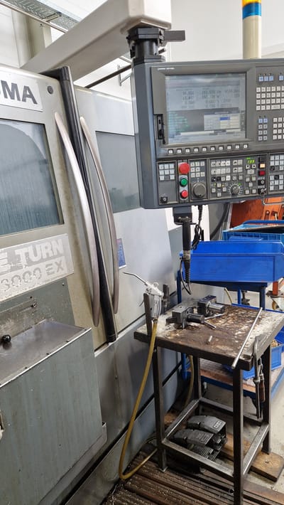This media file shows a close-up, eye-level shot of industrial machinery in a workshop. The primary focus is on a CNC (Computer Numerical Control) machine control panel and part of the machine itself. The control panel features a digital display screen showing various operational data, including "ACTUELLE POSITION" and numerical values. Below the screen are numerous buttons and a keypad, typical of industrial control systems.

To the right of the control panel, a metal fabrication or machining setup is visible, with a rough, weathered surface and various components. A yellow hose snakes across this area, likely for pneumatic or fluid delivery. In the foreground, several curved metal or plastic pieces are laid out, possibly workpieces or components related to the machinery.

The scene is indoors, in what appears to be a manufacturing or repair facility. The overall impression is one of an active workshop environment, though no people are visible in the frame. The lighting suggests it could be daytime, but the indoor setting makes it difficult to determine.

Visible text on the control panel includes "LOKUMA OSP-P200L", "WERKSTÜCKZÄHLER", and "RESTIG". Another prominent piece of text on the machine body reads "TURN 3000 EX". The location context is Ichenhausen, Germany. The image captures a detailed view of the operational interface and surrounding equipment of a complex industrial machine.
