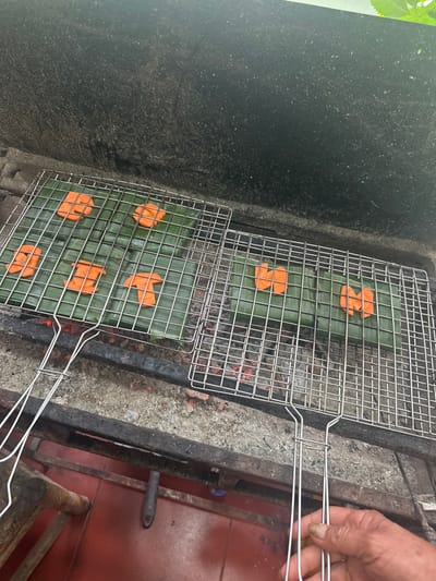 Two rectangular metal grill baskets sit over hot coals. Each holds several green banana leaves with orange, flower-shaped garnishes on top. A hand holding the grills is visible on the right side of the image. The grilling setup is on a concrete and metal surface, with a hint of a red tile floor visible in the lower right corner.
