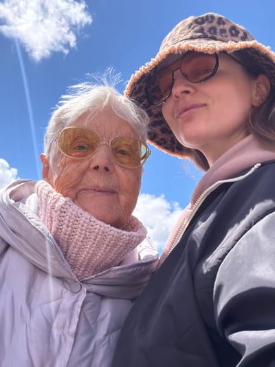The image shows two people, likely related, standing close together outdoors. The younger person wears sunglasses and a leopard print hood, with a dark jacket partially visible. The older person wears sunglasses and a light pink knitted scarf with a light-colored jacket. Both individuals face the camera from a low angle. The background consists of a blue sky with scattered white clouds. There is no indication of the city of Valozhyn, Belarus.