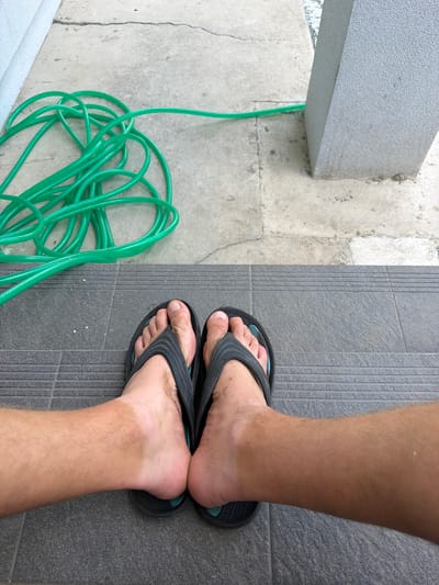 The image is a two-part composition. The top half shows a coiled green garden hose lying on a light-colored concrete surface. To the right, a grey rectangular pillar is visible. The bottom half of the image shows a pair of feet wearing black flip-flops, standing on dark grey tiled steps. The feet are lightly dusted with dirt. The scene appears to be an outdoor area, possibly a backyard or a patio, in the city of Bašť, Czechia.