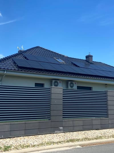 The image shows a close-up view of a modern building facade featuring a large array of solar panels installed on a dark tiled roof. The solar panels are arranged in neat rows, reflecting the bright blue sky. Adjacent to the solar panels, the building's exterior wall is covered with dark, vertical slats, interspersed with two outdoor air conditioning units, each bearing the LG logo. Below the slatted facade, a small window is visible. To the right of the building, a narrow strip of light-colored gravel borders a concrete curb and an asphalt road, suggesting an urban or suburban setting. A chimney with a metallic cap can be seen on the roof near the top left of the frame. The scene appears to be bathed in direct sunlight, indicating a clear, sunny day. No people are visible in the image. The location is Bolesławiec, Poland.