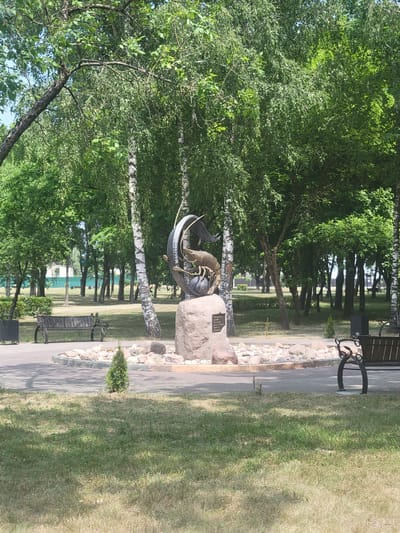 This media file depicts a park scene in Belaazyorsk, Belarus. The primary subject is a bronze sculpture of a crayfish perched on a rock. The sculpture is situated next to a paved pathway lined with decorative stones. Beyond the pathway, a well-maintained grassy area is visible, dotted with benches and trees. The background features a line of birch trees with their distinctive white bark, casting shadows on the grass. Further in the distance, more trees and some low buildings can be seen, suggesting a park or urban green space.
The scene appears to be set during the daytime, with bright sunlight creating distinct shadows, indicating a sunny day. The foliage is lush and green, suggesting it is springtime or summer. There are no people visible in the frame. The overall atmosphere is peaceful and serene.
A notable detail on the sculpture is a metal plaque attached to the rock base, which contains visible text. The text appears to be in a Cyrillic alphabet and reads "Macrobachium" followed by some other text that is difficult to decipher clearly. This likely identifies the species of crayfish depicted in the sculpture. The presence of the sculpture suggests it is a point of interest or a monument within the park.