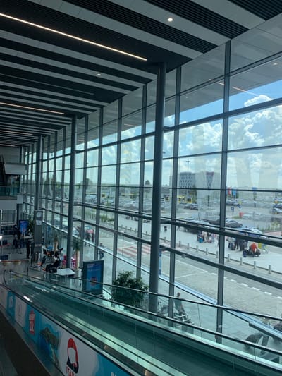 The image shows the interior of Katowice Airport, located in Pyrzowice, Poland. The airport is a modern building with large windows that offer a view of the outside. The image focuses on a downwards view of an escalator within the airport, situated beneath a glass wall. The escalator is made of metal and glass, and it descends from the upper level. The background features a view through the windows, showcasing a parking lot filled with cars and a distant building. A few people are visible in the distance, walking around the parking lot, and some people are seen near the escalator. There are also signs advertising various destinations, such as Italy. The image was captured on a sunny day with clear skies and is reminiscent of a busy airport environment.