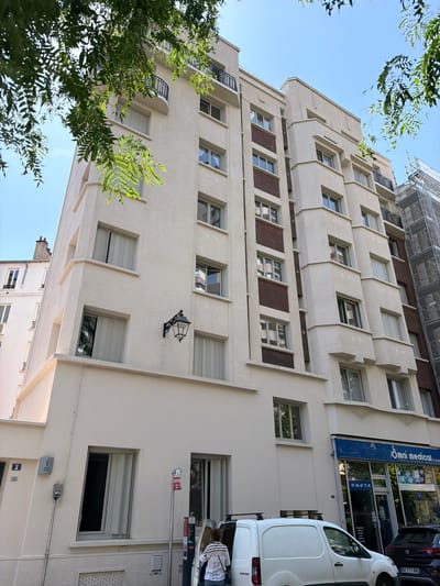 This is a vertical, eye-level shot of a cream-colored building with a modern architectural style. The building features multiple floors with numerous windows, many of which are equipped with horizontal blinds. Some windows are recessed, creating depth and shadow. The facade has textured plastering and decorative horizontal bands. A few sections of the building are adorned with dark brown brickwork.

In the foreground, lush green foliage from trees obscures parts of the lower and upper sections of the building, suggesting an outdoor setting with abundant plant life. The sky is clear and blue, indicating fair weather.

On the side of the building, a white commercial van is parked. Above the van, a storefront with a blue awning displays signage. The text on the awning reads "Omni medica" and "01 46 67 71 41". There are also other smaller signs with French text, and a license plate on a black car partially visible that reads "BZ-711-RH".

A vintage-style black lantern is mounted on the wall of the building, adding a decorative element. In the upper part of the image, scaffolding is visible, suggesting ongoing construction or renovation. The overall scene appears to be a street in Courbevoie, France, on a clear day. No people are clearly visible, but parts of individuals can be seen through windows and near the van. The image's orientation is rotated 90 degrees counter-clockwise.