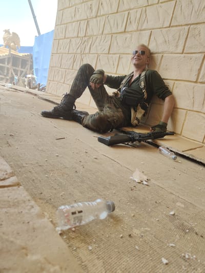 A man in military-style clothing is sitting against a wall. He is wearing sunglasses, a green shirt, camouflage pants, black boots, and gloves. A rifle and two plastic water bottles are on the ground near him. In the background, there is what appears to be a movie set with some people, construction materials, and a large statue of a mythological bird. The location is not specified in the image.