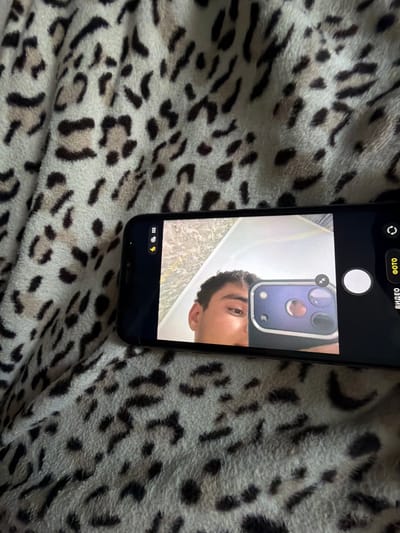 The image shows a person's face reflected in the camera of a smartphone. The phone is positioned on a leopard-print blanket. The person appears to be male, with dark hair and an eye visible in the reflection. The background of the reflection shows a light-colored wall and a mirror. The phone screen displays a camera interface with controls for photo and video modes. The text "ВИДЕО" (VIDEO) and "ФОТО" (PHOTO) are visible on the screen in Russian. The scene is indoors, with soft lighting. The overall mood is casual and personal, suggesting a selfie or a test shot. The location context provided is Avdarma, Moldova, but there are no specific visual cues in the image to confirm this location.